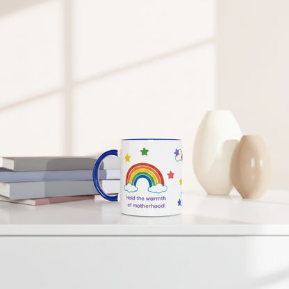 A white ceramic mug with a blue handle and rim, decorated with a colorful rainbow, stars, and the text 'Held the warmth of motherhood!', styled on a white surface with books and two vases in the background.