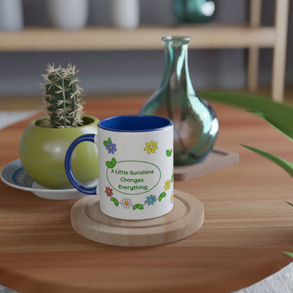 A white ceramic mug with a blue interior and handle, decorated with colorful flowers and the text 'A Little Sunshine Changes Everything', displayed on a wooden coaster on a wooden table.