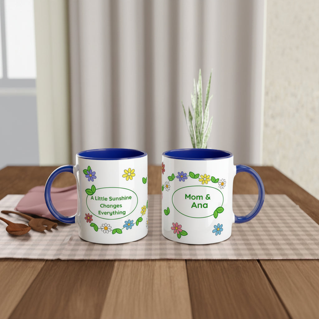 Two white ceramic mugs with blue interiors and handles are shown on a wooden table. One mug reads 'A Little Sunshine Changes Everything' with floral decoration, and the other reads 'Mom & Ana' in a similar floral design.