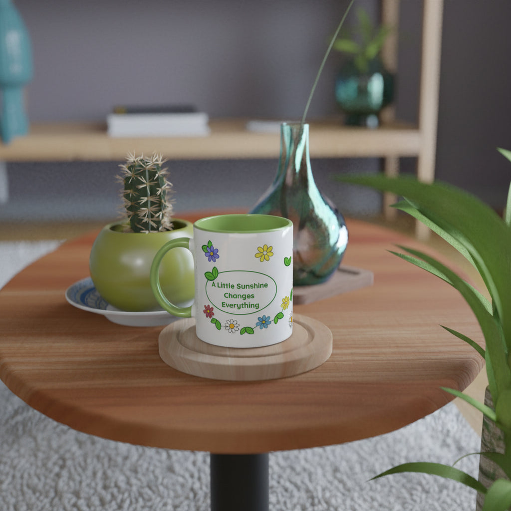 A white ceramic mug with a green handle and interior, decorated with colorful flowers and the phrase 'A Little Sunshine Changes Everything' inside a green oval. The mug sits on a round wooden coaster on a round wooden table, styled with a small potted cactus and a tall green glass vase in a modern living room setting.