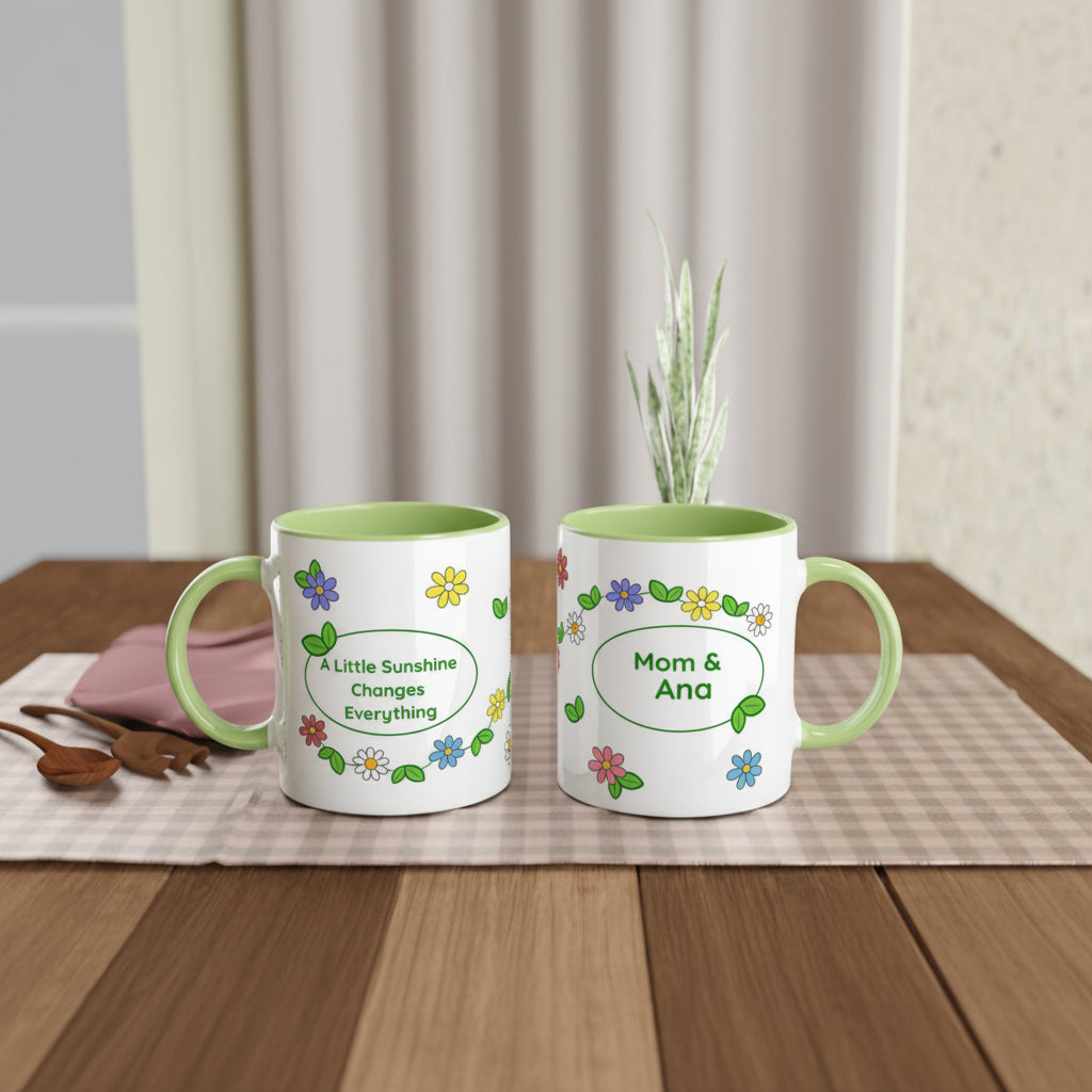 Two white ceramic mugs with light green interiors and handles are shown on a wooden table. One mug has the phrase 'A Little Sunshine Changes Everything' with small colorful flowers, and the other says 'Mom & Ana' in a similar floral design.