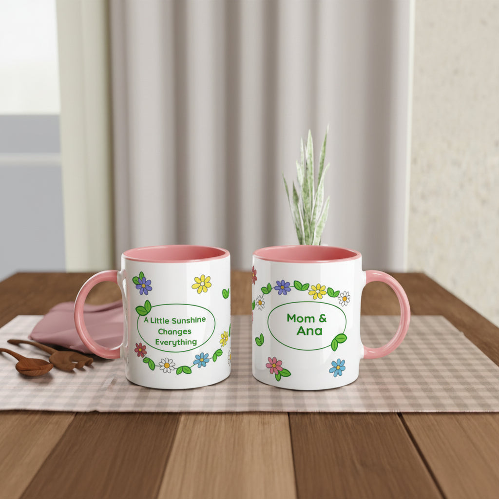 Two white ceramic mugs with pink interiors and pink handles sit on a wooden table. The left mug reads 'A Little Sunshine Changes Everything' with small colorful flowers, and the right mug reads 'Mom & Ana' with matching floral decoration.