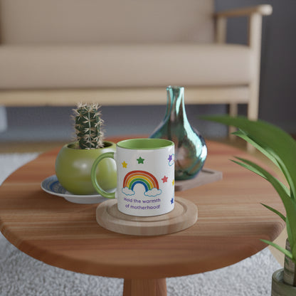 A white ceramic mug with a green handle and interior, printed with a colorful rainbow, stars, and the phrase 'hold the warmth of motherhood', sits on a wooden coaster on a round wooden table in a styled living room setting.