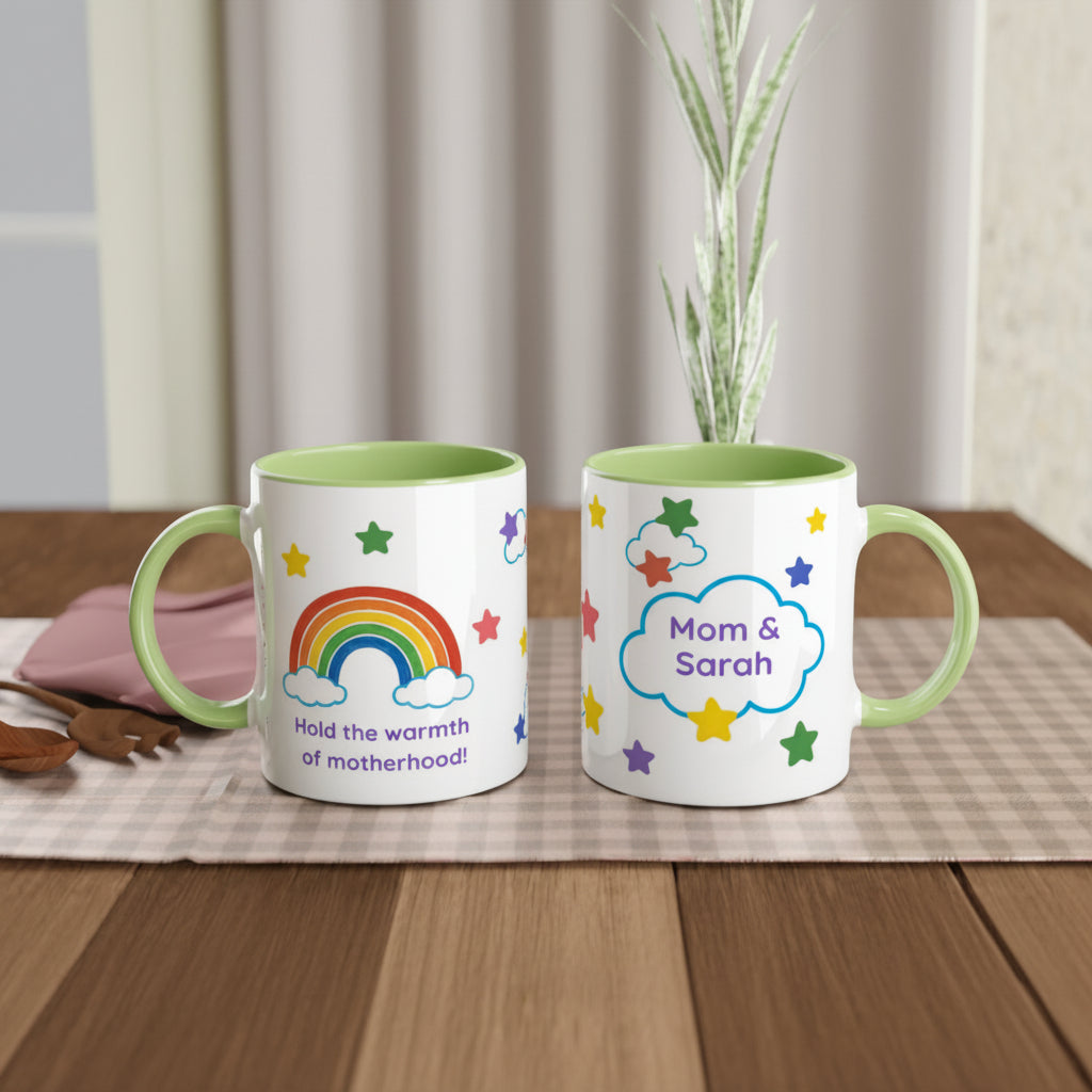 Two white ceramic mugs with light green interiors and handles sit on a wooden table. One mug shows a rainbow, clouds, stars, and the text 'Hold the warmth of motherhood!'.