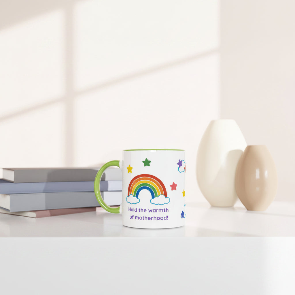 A white ceramic mug with a green handle and rim, decorated with a colorful rainbow, stars, and the text 'Hold the warmth of motherhood!!' in purple. The mug is styled on a white surface next to stacked books and two minimalist ceramic vases under soft window-light shadows.