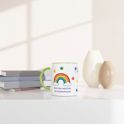 A white ceramic mug with a green handle and rim, decorated with a colorful rainbow, stars, and the text 'Hold the warmth of motherhood!!' in purple. The mug is styled on a white surface next to stacked books and two minimalist ceramic vases under soft window-light shadows.