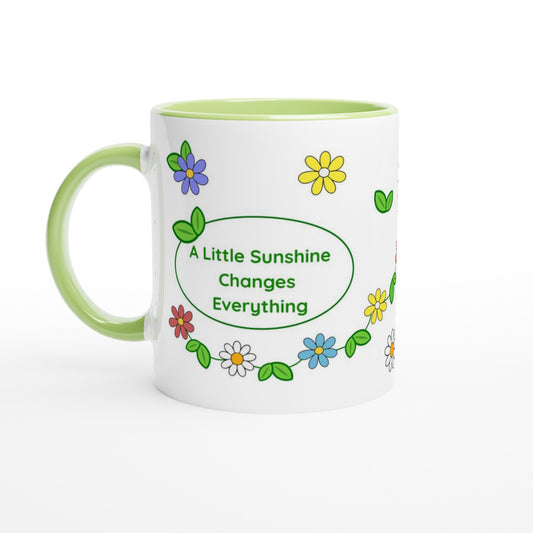 A white ceramic mug with a light green handle and interior, decorated with colorful cartoon flowers and a green oval that reads 'A Little Sunshine Changes Everything'.