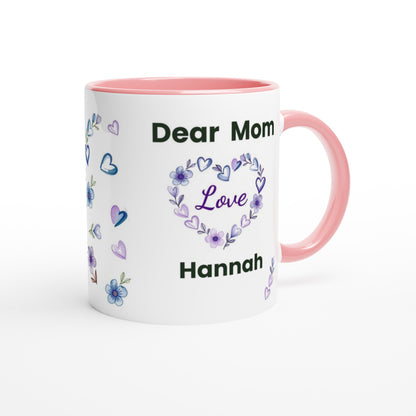 A white ceramic mug with a pink interior and handle, decorated with purple and blue floral heart designs and the text 'Dear Mom Love Hannah'.