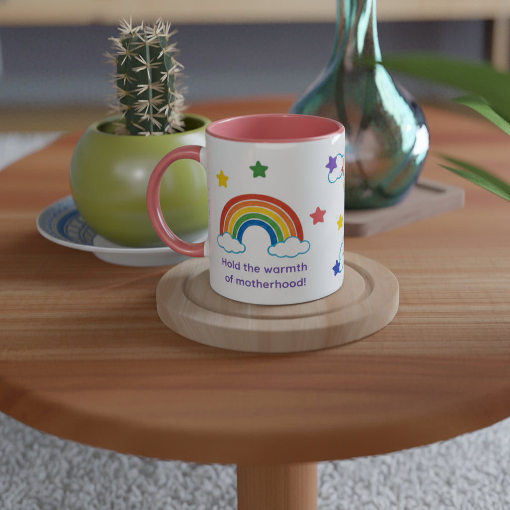 A white ceramic mug with a pink handle and interior, decorated with a colorful rainbow, stars, clouds, and the text 'Hold the warmth of motherhood!'. The mug sits on a round wooden coaster on a wooden table, with a potted cactus and a glass vase in the softly blurred background.