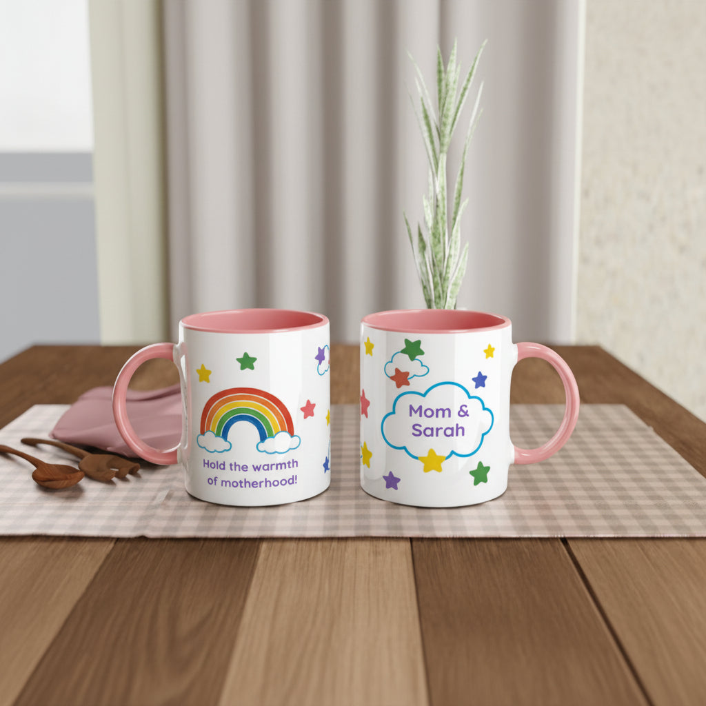 Two white ceramic mugs with pink interiors and handles are displayed on a wooden table. One mug features a colorful rainbow and the text 'Hold the warmth of motherhood', while the other has a cloud with the names 'Mom & Sarah' surrounded by stars.