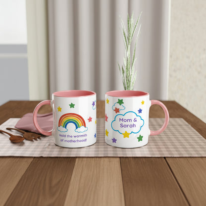 Two white ceramic mugs with pink interiors and handles are displayed on a wooden table. One mug features a colorful rainbow and the text 'Hold the warmth of motherhood', while the other has a cloud with the names 'Mom & Sarah' surrounded by stars.