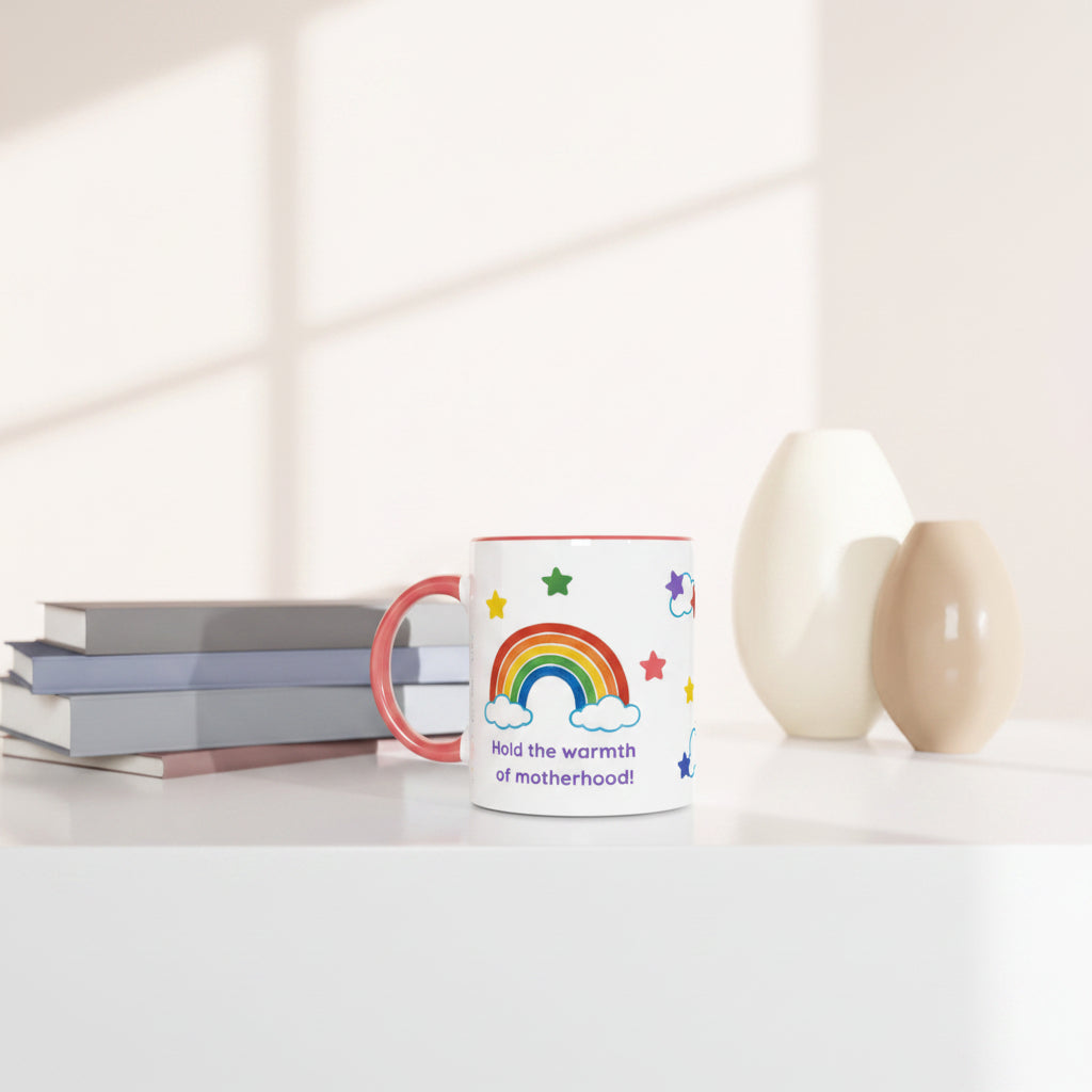 A white ceramic mug with a colorful rainbow, stars, and the text 'Hold the warmth of motherhood!' sits on a white tabletop next to stacked books and two matte vases.