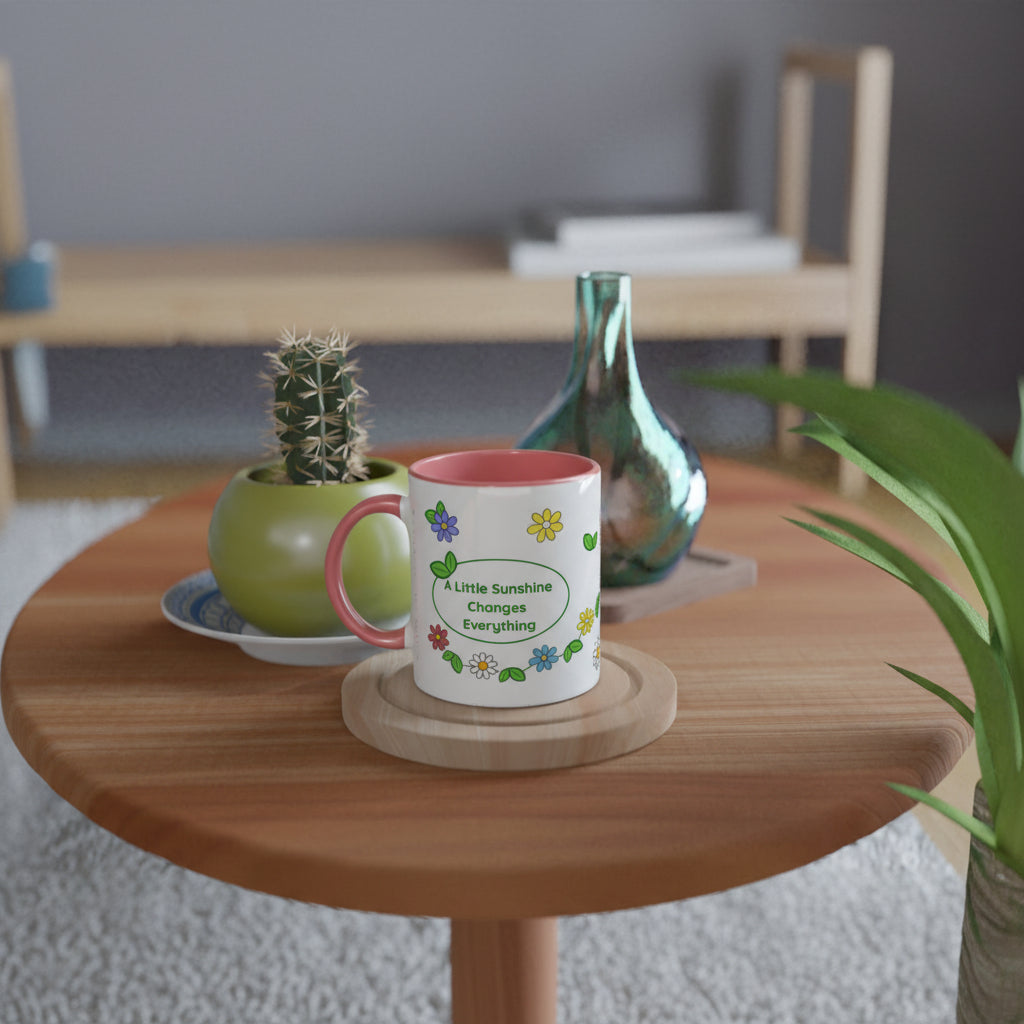 A white ceramic mug with a pink interior and handle, printed with the phrase 'A Little Sunshine Changes Everything' in green text surrounded by colorful flowers, sitting on a wooden coaster on a round wooden table.