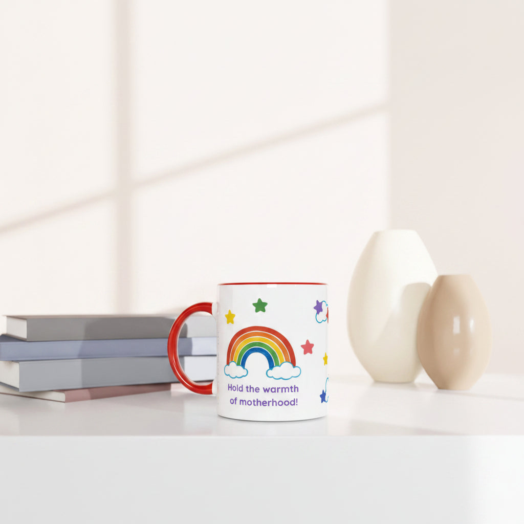 A white ceramic mug with a red handle and rim, decorated with a colorful rainbow, stars, and the text 'Hold the warmth of motherhood!'. The mug is styled on a white surface next to stacked books and two ceramic vases in a bright, minimalist room.