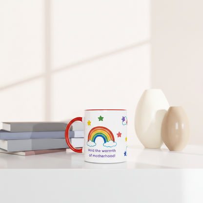 A white ceramic mug with a red handle and rim, decorated with a colorful rainbow, stars, and the text 'Hold the warmth of motherhood!'. The mug is styled on a white surface next to stacked books and two ceramic vases in a bright, minimalist room.