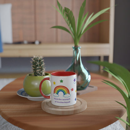 A white ceramic mug with a red handle and interior, decorated with a colorful rainbow, stars, and the text 'Hold the warmth of motherhood!', displayed on a wooden coaster on a round wooden table.