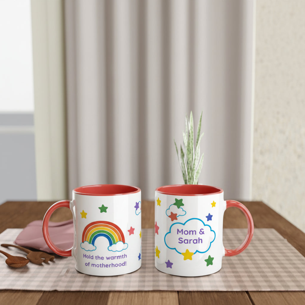 Two white ceramic mugs with coral-red interiors and handles are displayed on a wooden table. One mug features a colorful rainbow and the text 'Hold the warmth of motherhood!', while the other has a cloud with 'Mom & Sarah' surrounded by stars.
