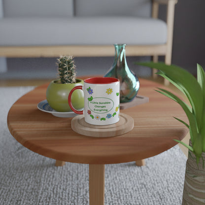 A white ceramic mug with a red handle and interior sits on a round wooden coaster on a round wooden table. The mug is decorated with colorful flowers and a speech bubble that reads 'A Little Sunshine Changes Everything'.