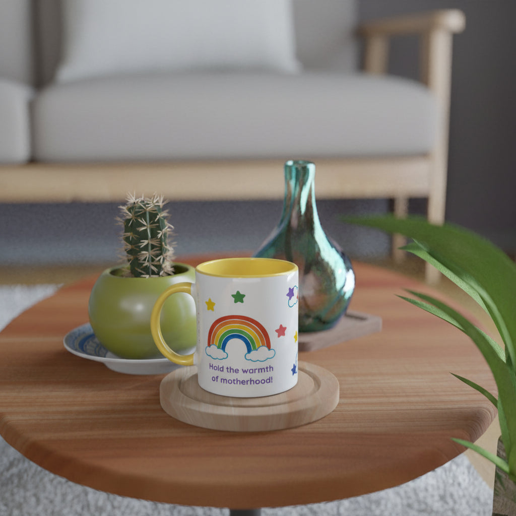 A white ceramic mug with a yellow handle and interior, printed with a colorful rainbow, stars, and the phrase 'had the warmth of motherhood', displayed on a round wooden table in a cozy living room setting.