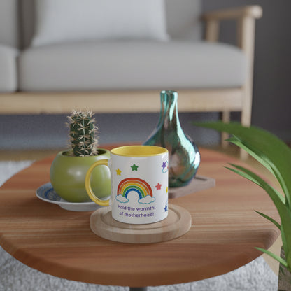 A white ceramic mug with a yellow handle and interior, printed with a colorful rainbow, stars, and the phrase 'had the warmth of motherhood', displayed on a round wooden table in a cozy living room setting.
