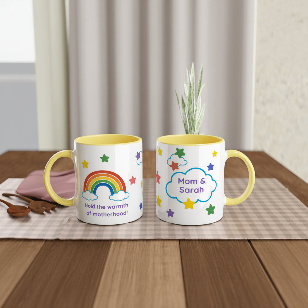 Two white ceramic mugs with yellow interiors and handles are displayed side by side on a wooden table. The left mug features a colorful rainbow, clouds, stars, and the text 'Hold the warmth of motherhood!'.