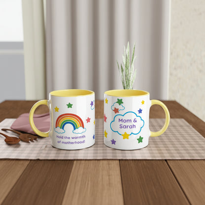 Two white ceramic mugs with yellow interiors and handles are displayed side by side on a wooden table. The left mug features a colorful rainbow, clouds, stars, and the text 'Hold the warmth of motherhood!'.