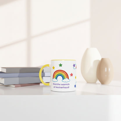 A white ceramic mug with a yellow handle and rim, decorated with a colorful rainbow, stars, clouds, and the text 'Hold the warmth of motherhood!', styled on a white table next to books and two ceramic vases.