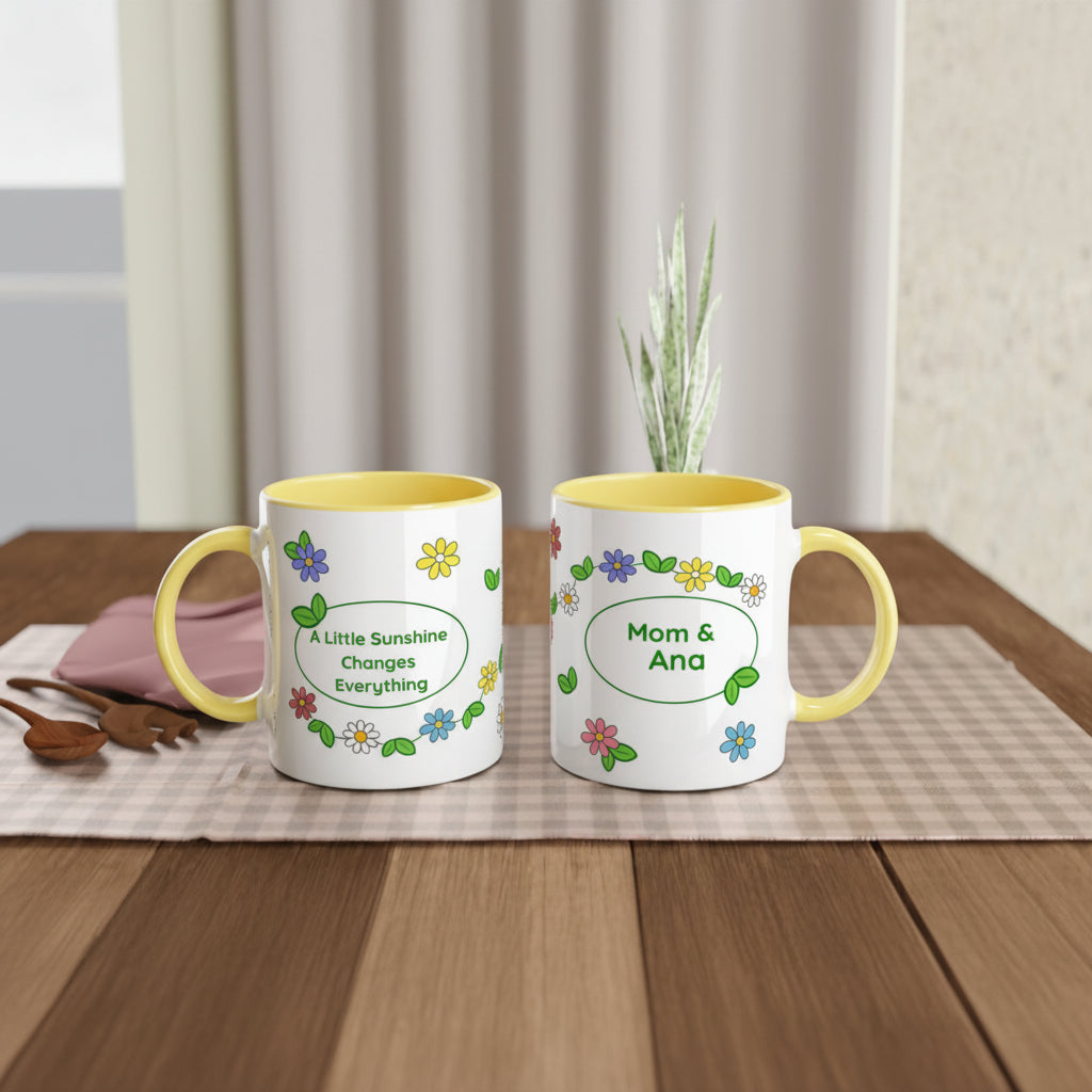 Two white ceramic mugs with yellow interiors and handles, decorated with colorful flowers and green leaves, displayed on a wooden table. One mug reads 'A Little Sunshine Changes Everything' and the other reads 'Mom & Ana'.