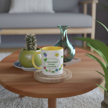 A white ceramic mug with a yellow handle and interior sits on a wooden coaster on a round wooden table. The mug is decorated with colorful flowers and the text 'A Little Sunshine Changes Everything'.