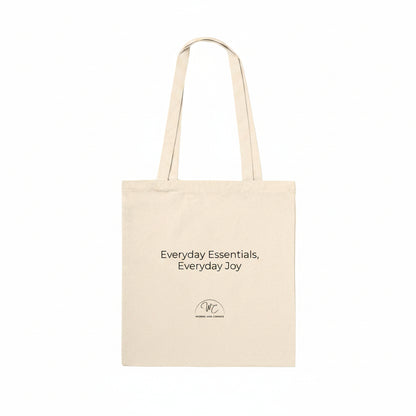 A natural beige canvas tote bag with black printed text reading 'Everyday Essentials, Everyday Joy' and a small circular logo below, shown on a plain white background.