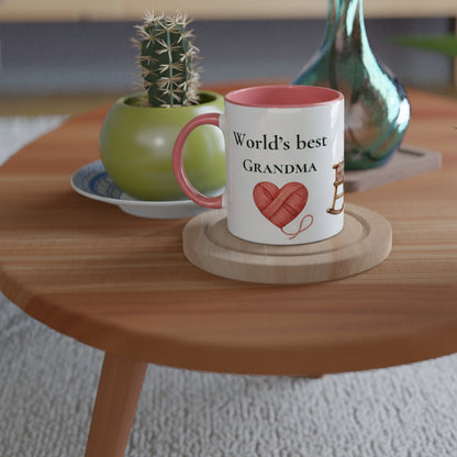 A white ceramic mug with a pink interior and handle sits on a round wooden coaster on a light wood table. The mug is printed with the text 'World's best GRANDMA' and a red heart made of yarn