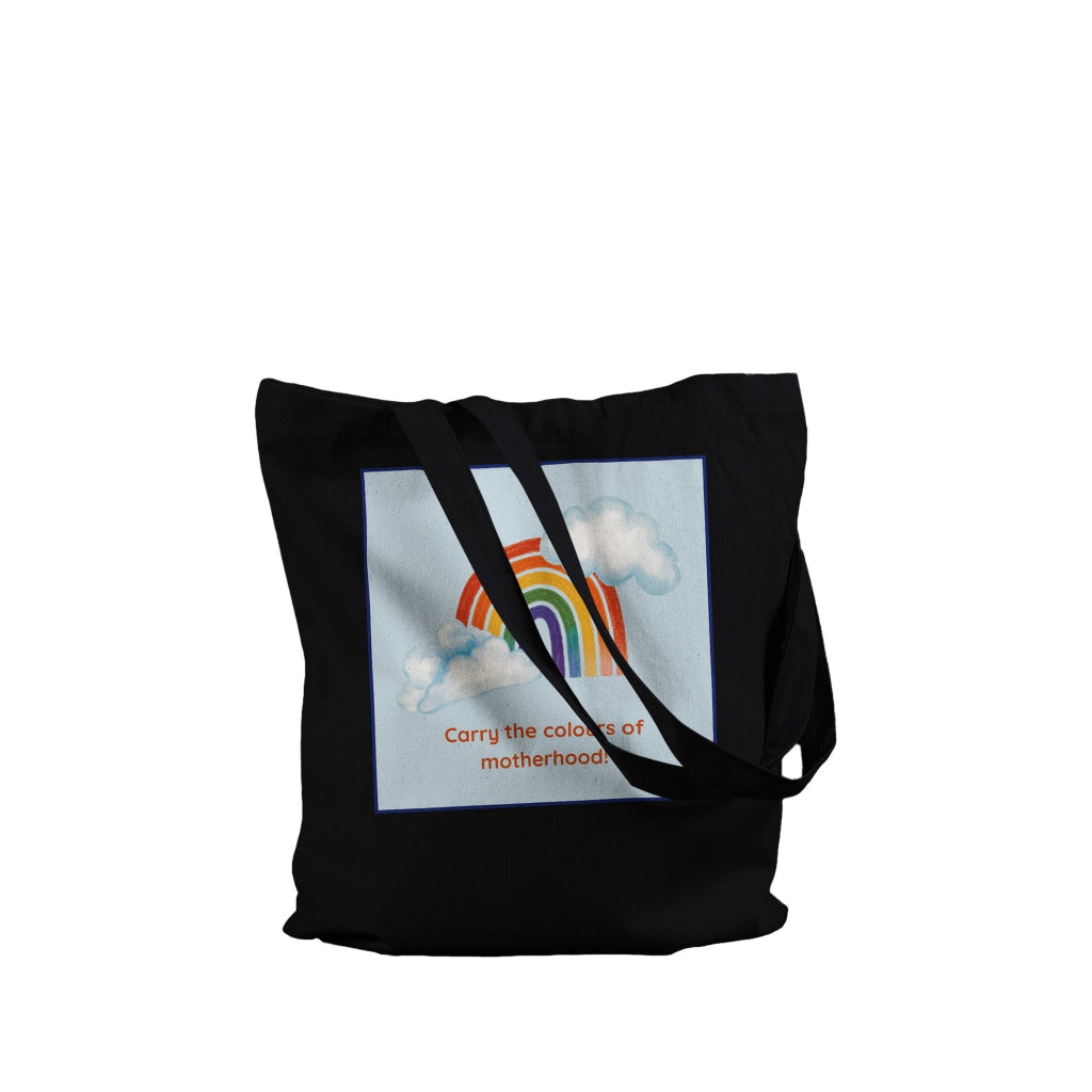 A black canvas tote bag with long shoulder straps, featuring a square graphic print on the front. The print shows a pastel rainbow arching over white clouds on a light blue background, with the text 'Carry the colors of motherhood' printed below in orange.