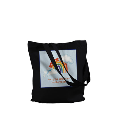 A black canvas tote bag with long shoulder straps, featuring a square graphic print on the front. The print shows a pastel rainbow arching over white clouds on a light blue background, with the text 'Carry the colors of motherhood' printed below in orange.