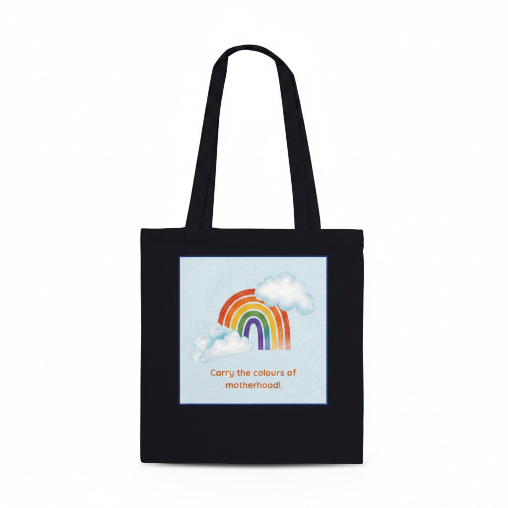A black canvas tote bag with two shoulder straps, featuring a square graphic print on the front. The print shows a pastel rainbow arching over white clouds on a light blue background, with the text 'Carry the colours of motherhood!' printed below in orange.