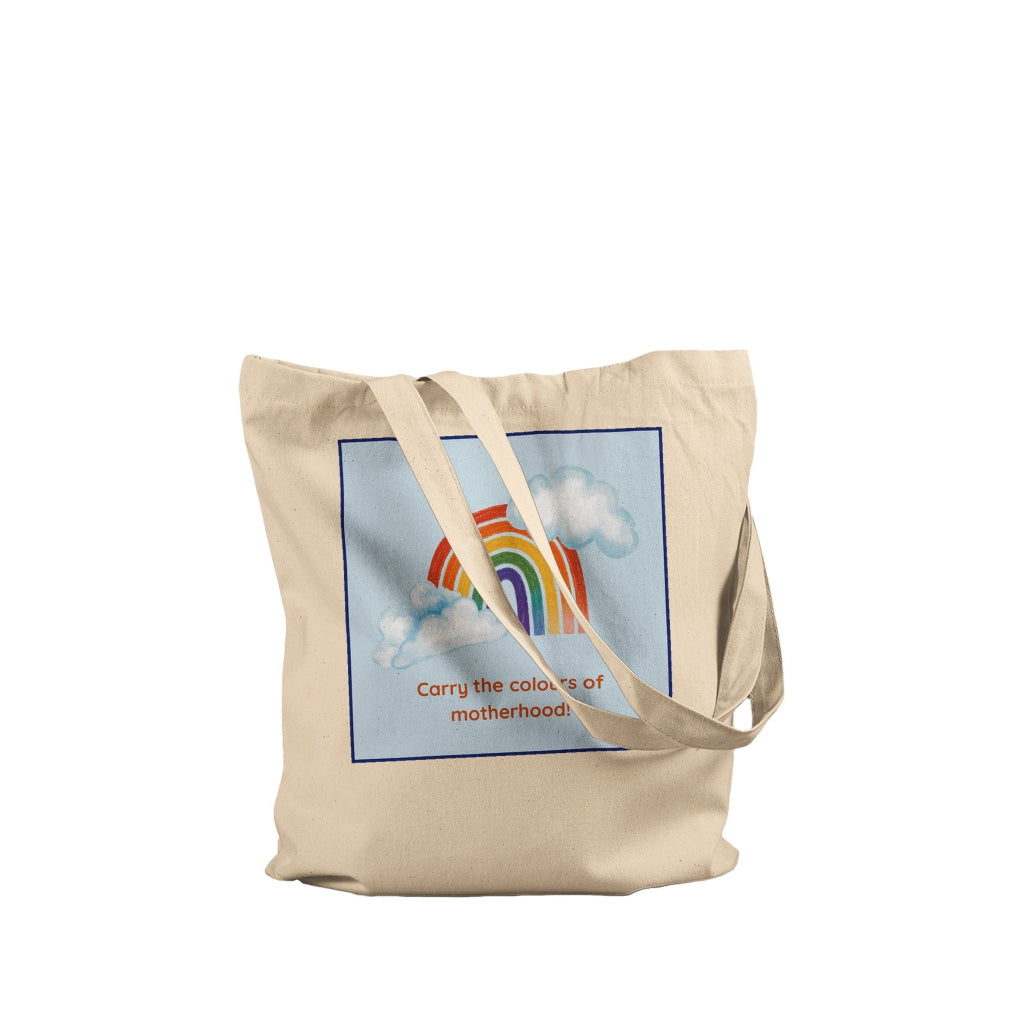 A beige canvas tote bag with a printed graphic of a rainbow and clouds on a light blue background, with the text 'Carry the colours of motherhood' written below. The bag is shown against a plain white background.
