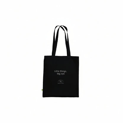 A black canvas tote bag with white printed text reading 'Little things, big joys' and a small circular logo near the bottom, shown against a plain white background.