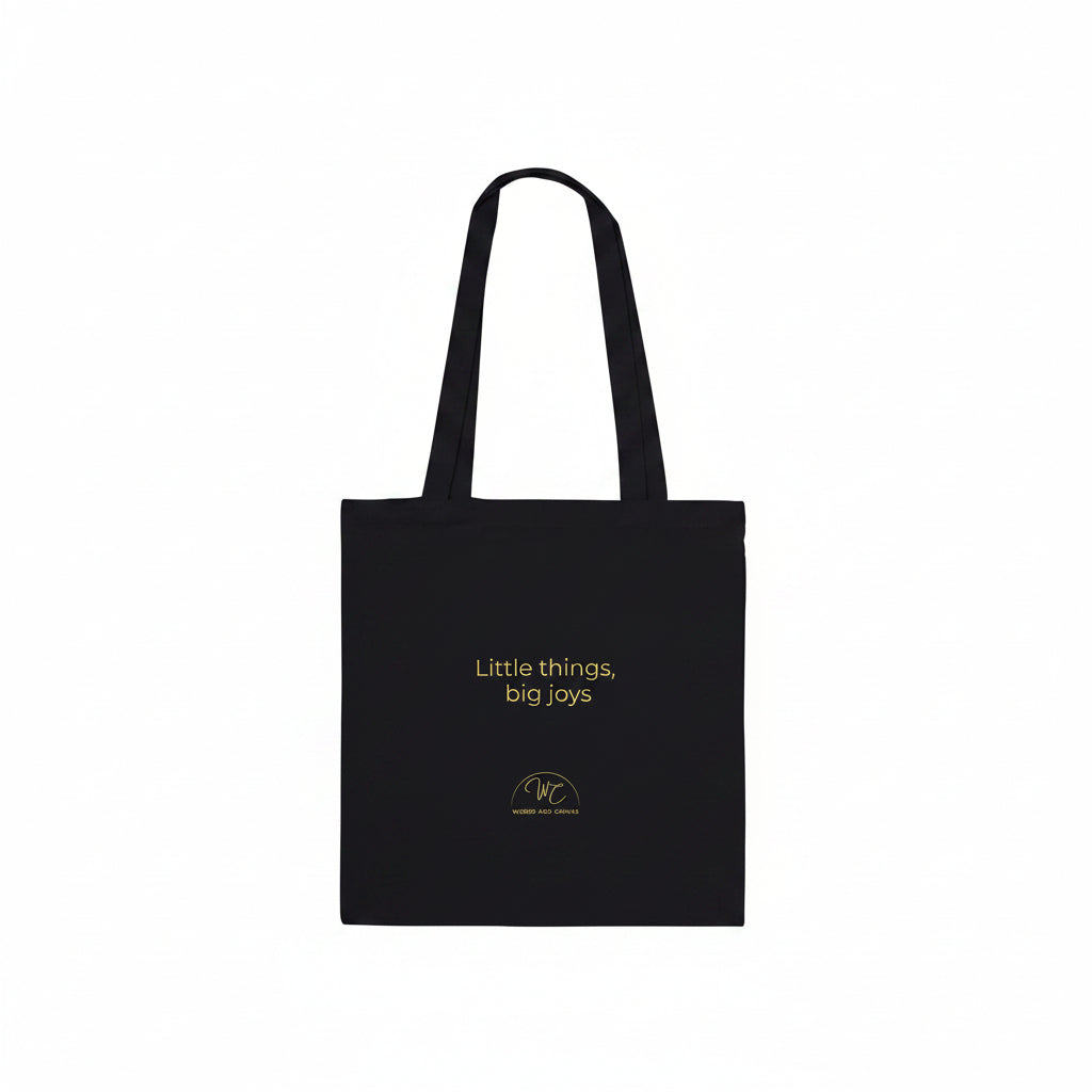 A black canvas tote bag with two shoulder straps, featuring the text 'Little things, big joys' and a small circular logo printed in yellow , set against a plain white background.
