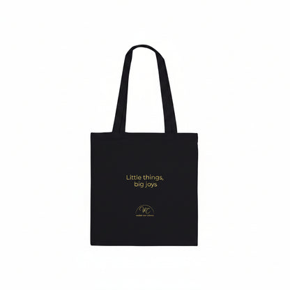 A black canvas tote bag with two shoulder straps, featuring the text 'Little things, big joys' and a small circular logo printed in yellow , set against a plain white background.