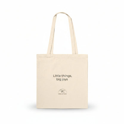 A natural canvas tote bag with black printed text reading 'Little things, big joys' and a small circular logo below, shown on a clean white background.