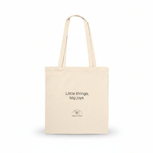 A natural canvas tote bag with black printed text reading 'Little things, big joys' and a small circular logo below, shown on a clean white background.