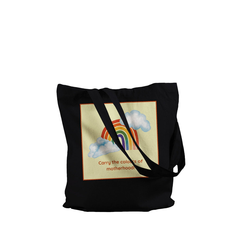 A black canvas tote bag with long shoulder straps, featuring a square graphic print on the front. The print shows a pastel rainbow arching over light blue clouds on a cream background, with the text 'Carry the colors of motherhood' in a reddish-brown font.