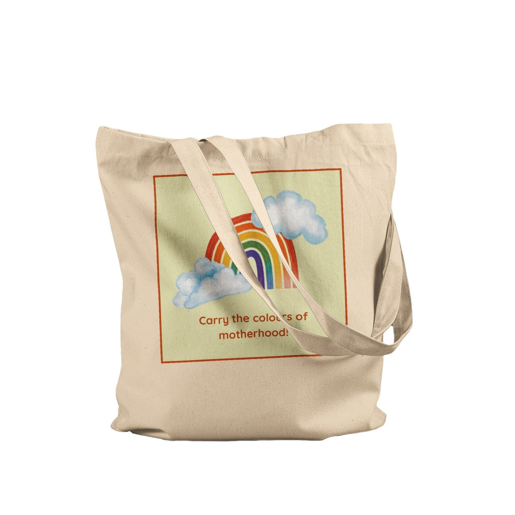 A beige canvas tote bag with a printed graphic of a rainbow and clouds inside a red border, with the text 'Carry the colours of motherhood'.