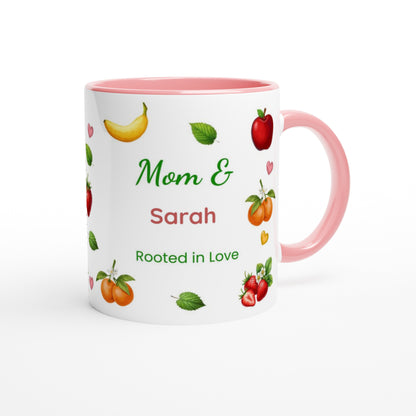 A white ceramic mug with a pink interior and handle, decorated with illustrations of fruits and the personalized text 'Mom & Sarah Rooted in Love'.