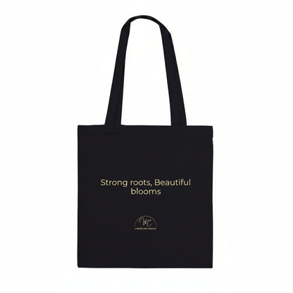 A black canvas tote bag with two shoulder straps, featuring the text 'Strong roots, Beautiful blooms' 