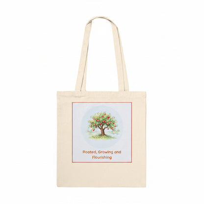A natural beige canvas tote bag with two shoulder straps, featuring a square graphic print on the front. The print shows a colorful watercolor-style tree with green leaves and red fruit inside a light blue circle, bordered by a thin red line, with the text 'Rooted, Growing and Flourishing' printed below in red.