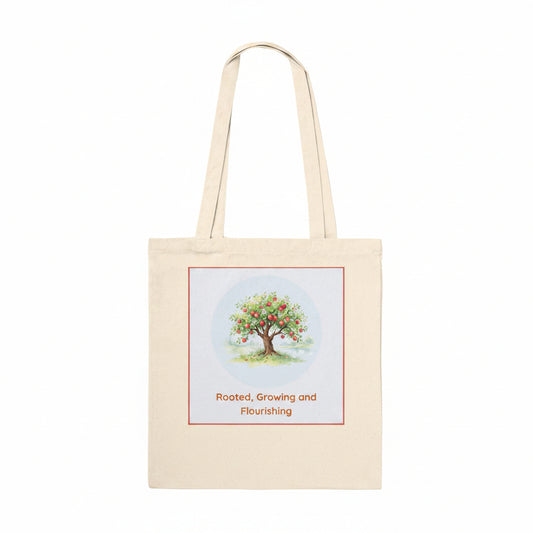 A natural beige canvas tote bag with two shoulder straps, featuring a square graphic print on the front. The print shows a colorful watercolor-style tree with green leaves and red fruit inside a light blue circle, bordered by a thin red line, with the text 'Rooted, Growing and Flourishing' printed below in red.