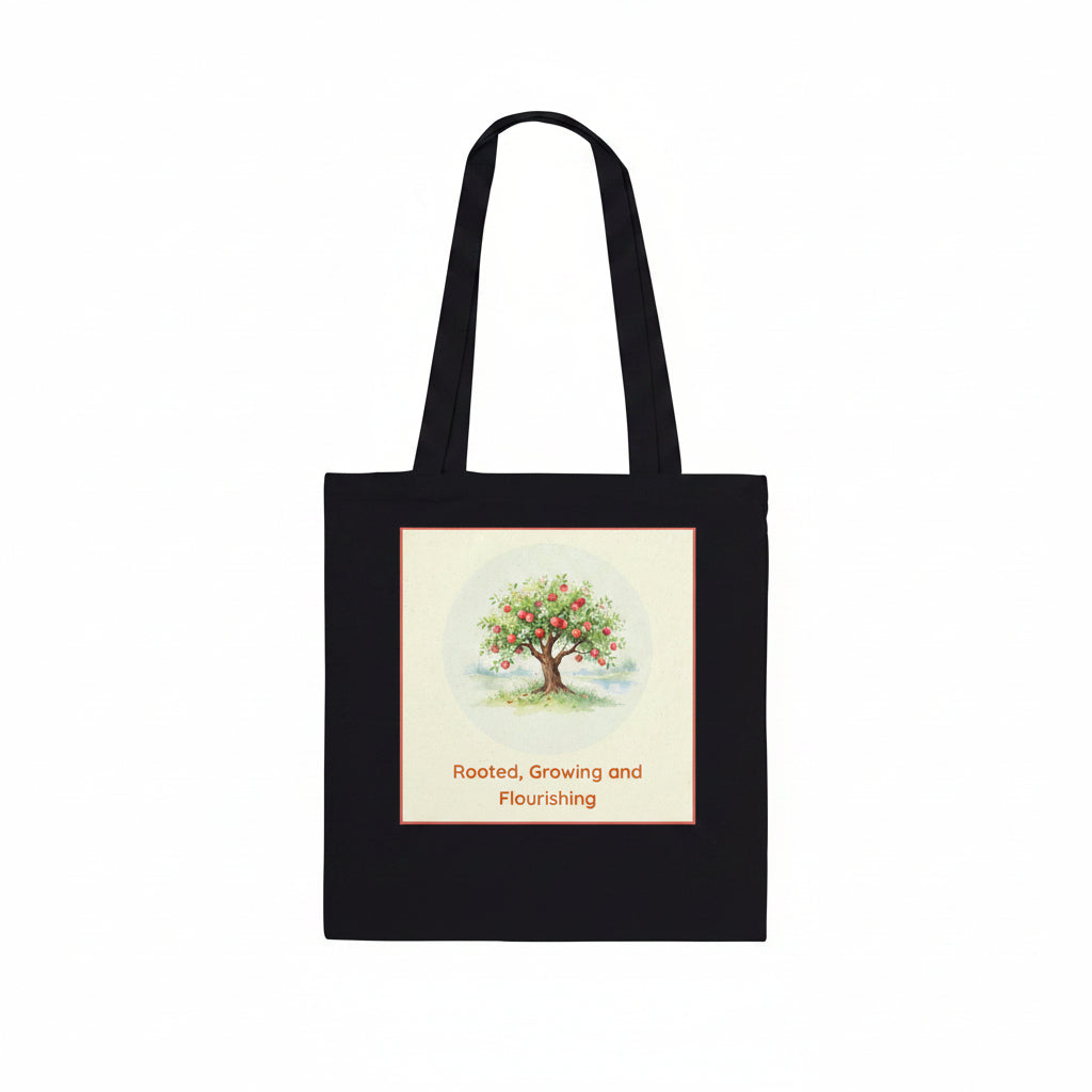 A black canvas tote bag with two shoulder straps, featuring a square graphic print on the front. The print shows a watercolor-style illustration of a tree with green leaves and red fruit, surrounded by a light cream border, with the text 'Rooted, Growing and Flourishing' printed below in red.