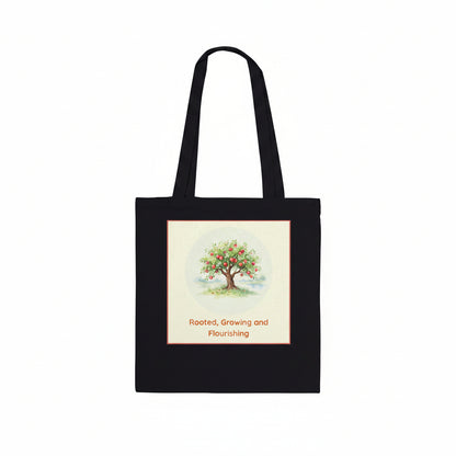 A black canvas tote bag with two shoulder straps, featuring a square graphic print on the front. The print shows a watercolor-style illustration of a tree with green leaves and red fruit, surrounded by a light cream border, with the text 'Rooted, Growing and Flourishing' printed below in red.