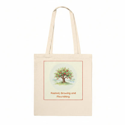 A natural canvas tote bag with two shoulder straps, featuring a printed graphic of a fruit tree with red apples and the text 'Rooted, Growing and Flourishing' on the front.