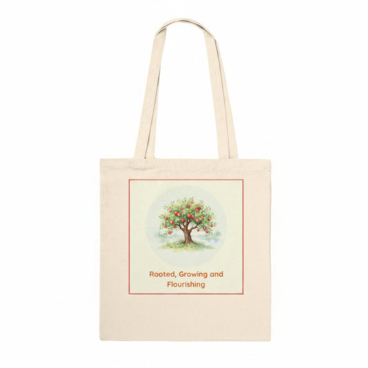 A natural canvas tote bag with two shoulder straps, featuring a printed graphic of a fruit tree with red apples and the text 'Rooted, Growing and Flourishing' on the front.